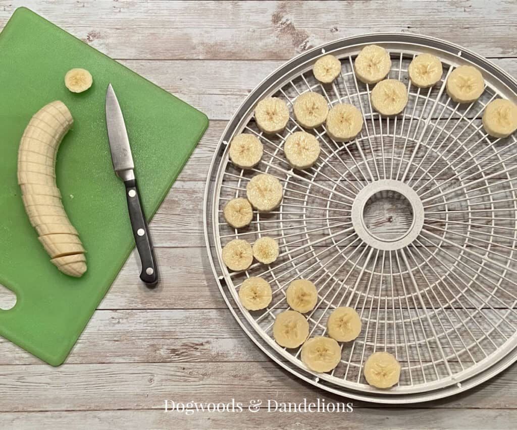 sliced bananas on a dehydrator tray beside a cutting board with a sliced banana and a knife