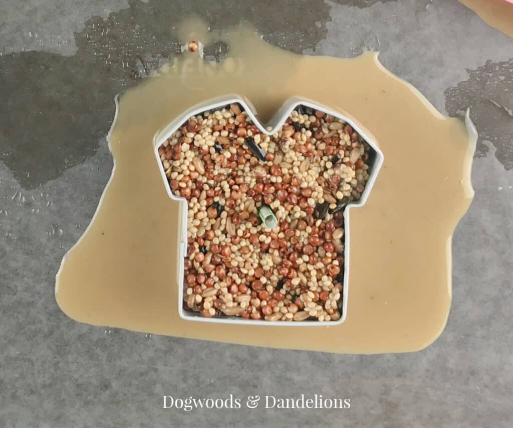 a coconut oil birdseed ornament in a tshirt shaped cookie cutter with peanut butter and coconut oil seeping our of it.