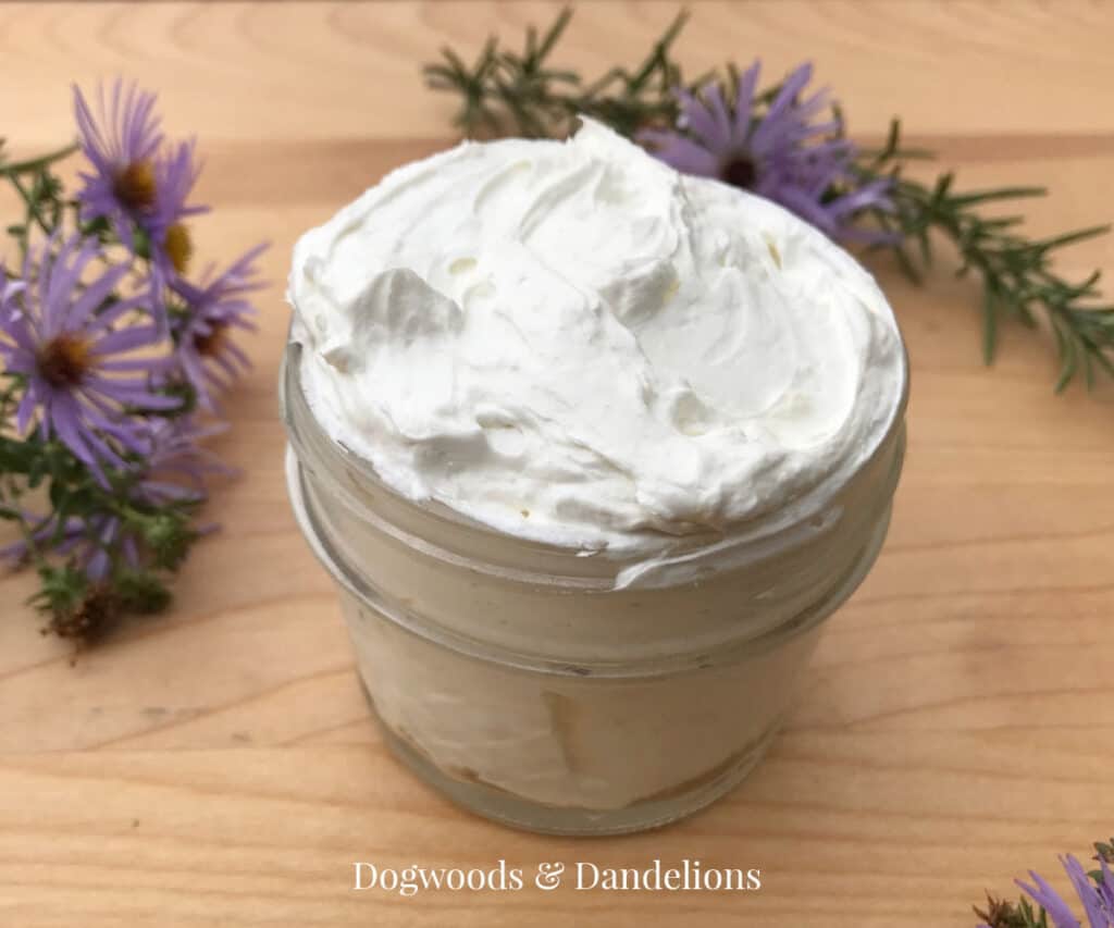 a jar of whipped body butter in front of rosemary springs and aster flowers.