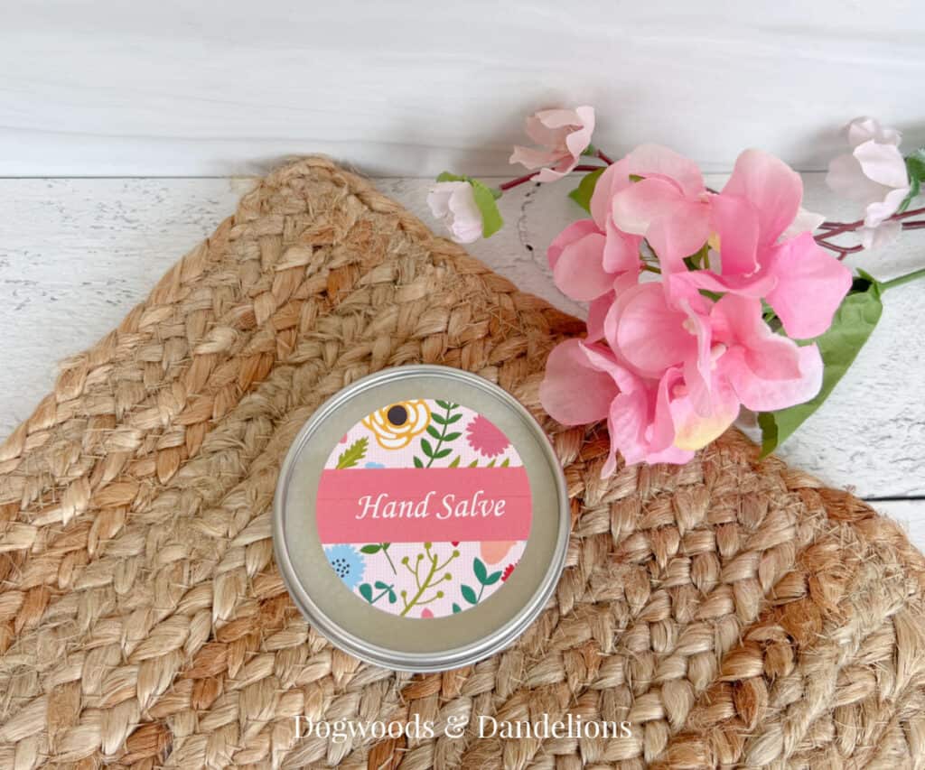 a tin of hand salve on a brown woven mat beside pink flowers.