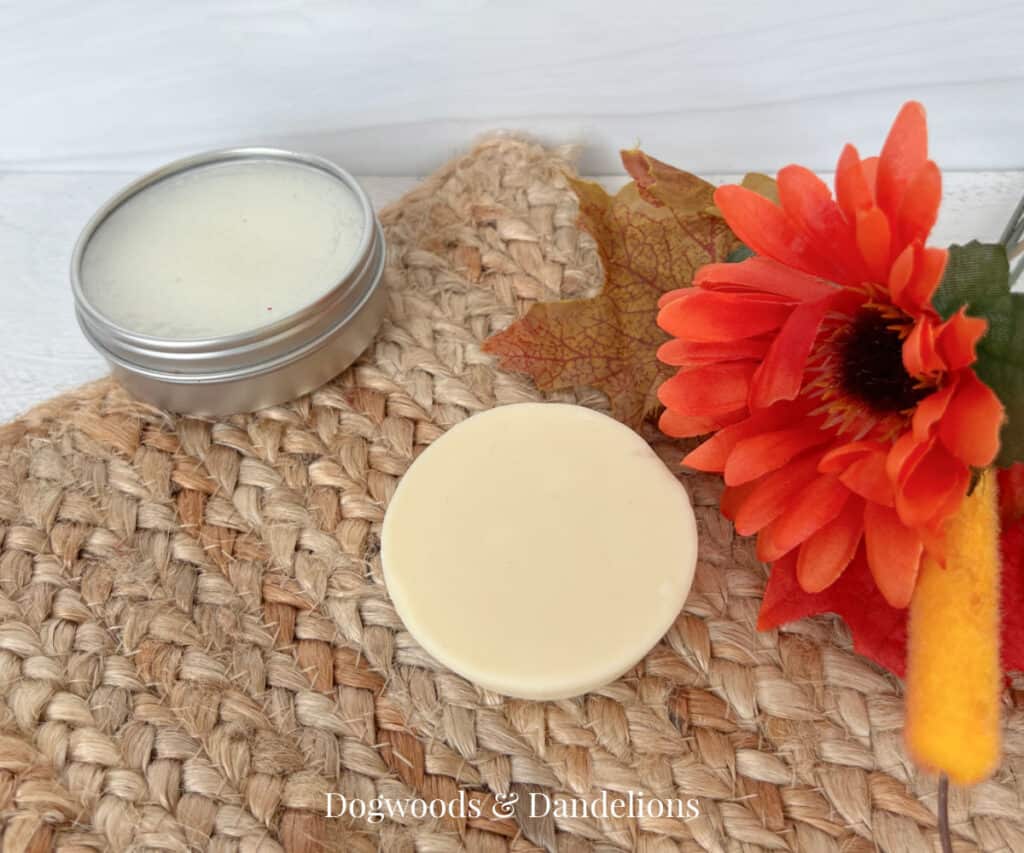 a lotion bar and a lotion bar in a tin on a brown woven mat beside an orange flower and leaves.