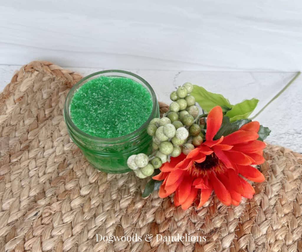 a jar of green sugar scrub beside an arrangement of flowers.