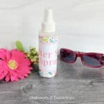 a bottle of after sun spray beside a flower, greenery, and a pair of sunglasses