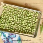 sliced okra on a sheet pan ready to be frozen beside a kitchen towel and okra pods