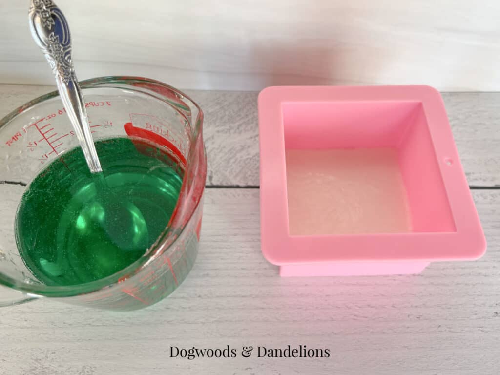 green soap base in a pyrex container with a spoon and white soap base in a pink mold